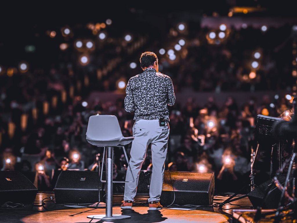 pexels photo 31129059 Speaker addressing large audience with phones lit up at night event.